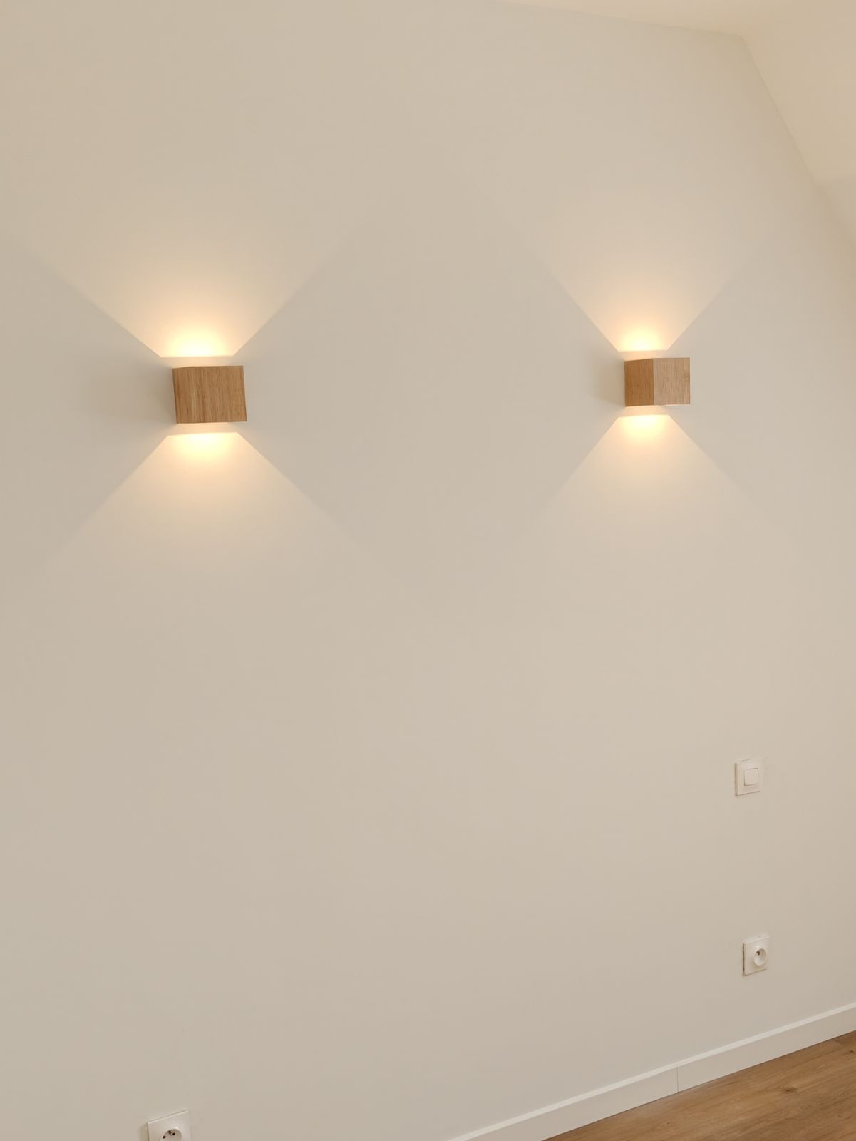 Deux appliques murales cubiques en bois, diffusant une lumière vers le haut et le bas, de part et d'autre d'un lit